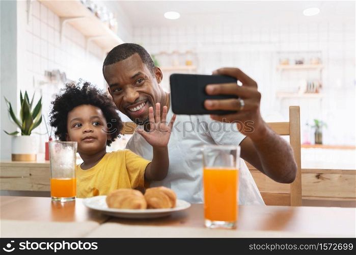 Happy African-American man and kid boy using mobile phone for taking selfie together at home. Cheerful African Father and son doing video chat and waving hand during talking to family with smartphone
