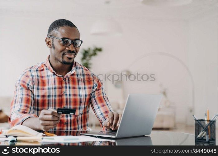 Happy african american man in glasses is purchasing online from home using laptop and credit card. Internet banking and money transfer. Ecommerce and consumerism concept.. Happy african american man in glasses is purchasing online using laptop and credit card.