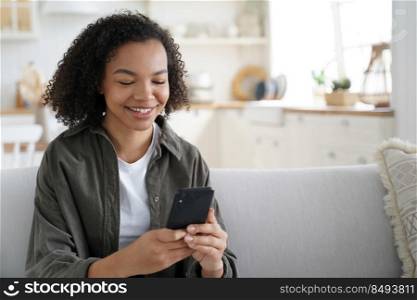 Happy african american teen girl share good news on social network via mobile phone sitting on sofa at home. Smiling mixed race young female enjoying shopping online using smartphone apps.. Happy african american teen girl holds smartphone using social network apps, shopping online at home