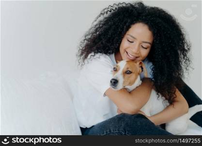 Happy African American woman expresses love to dog, embraces pet, dressed in casual wear, sit on comfortable bed in bedroom, sleeps with domestic animal. Jack russell terrier with owner in morning