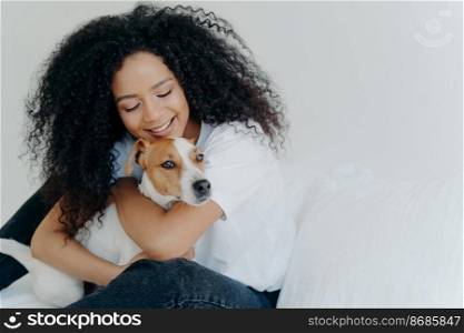 Happy African American woman expresses love to dog, embraces pet, dressed in casual wear, sit on comfortable bed in bedroom, sleeps with domestic animal. Jack russell terrier with owner in morning