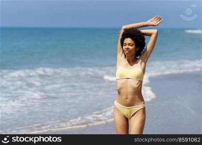 Happy African American woman raising her arms on the beach to enjoy her holiday in the sun. Young black female wearing yellow bikini.. Happy African American woman raising her arms on the beach to enjoy her holiday in the sun.