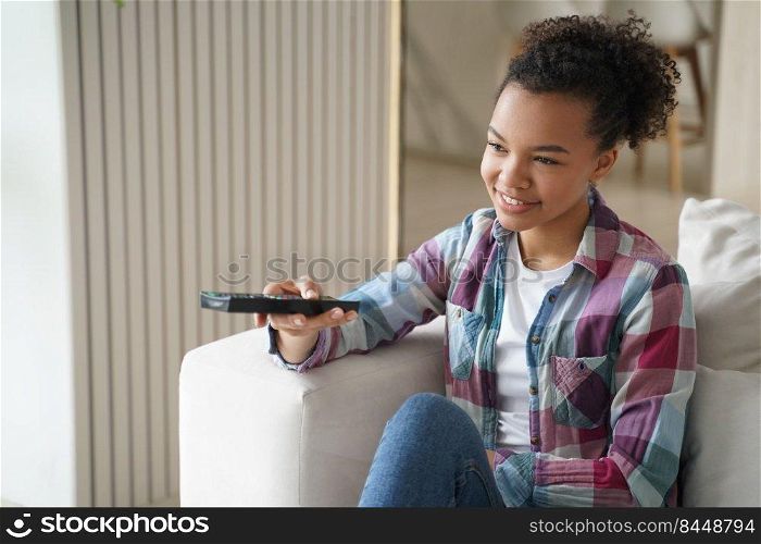 Happy african american young girl holding using tv controller, sitting on couch in living room, watching movie or series at home. Smiling teen lady relax at home spending leisure time with television.. African american young girl watching movie or tv series, enjoying television program at home