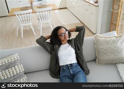 Happy african american young girl relaxing on comfortable couch in cozy living room at home. Biracial woman sitting on sofa with hands behind head, dreaming, enjoying weekend break.. Happy african american young girl relaxing on comfortable couch in cozy living room, enjoying break