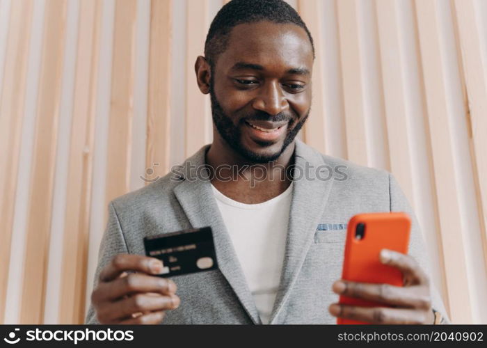 Happy african businessman in suit holding credit card and mobile phone while buying goods or services via Internet, smiling afro american man using payment e-banking system at work. Happy african businessman in suit holding credit card and mobile phone while doing online shopping