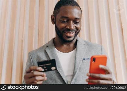 Happy african businessman in suit holding credit card and mobile phone while buying goods or services via Internet, smiling afro american man using payment e-banking system at work. Happy african businessman in suit holding credit card and mobile phone while doing online shopping