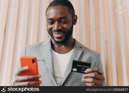 Happy african businessman in suit holding credit card and mobile phone while buying goods or services via Internet, smiling afro american man using payment e-banking system at work. Happy african businessman in suit holding credit card and mobile phone while doing online shopping