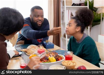 Happy African family father feeding food with daughter in dinner on festive thanksgiving eve day together at home, father and mother and children celebration party in xmas and new year in holiday.