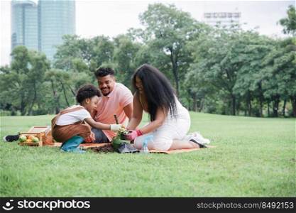 Happy African family picnic and doing activities together in park on vacation. Selective focus on father looking at Little son and his young mother wear gloves and plant a plant or flower in pot