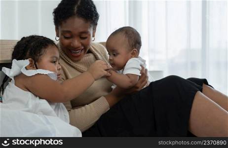Happy African family, young mother and daughters, little kid older sister looking and tender holding her newborn sister hand while mom looking at children with love and care at bedroom at home