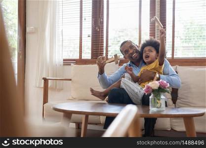 Happy African Father and little child son spending time playing wooden airplane toy at home together. Smiling Dad in blue shirt embracing kid boy in yellow casual after arriving from business working on couch in living room. Having fun. Black family concept.