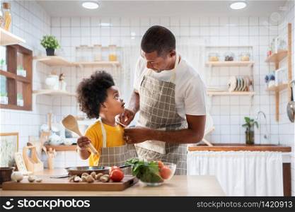 Happy African father and son dress up together before cooking in the white kitchen. Single Dad Chef with black kid helper in yellow shirt preparing food and looking at Each Other at home. Eye Contact, Relationship, Talking, Warm Family.