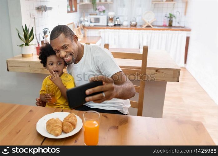 Happy African Father and son taking selfie photo with smartphone during lunch time in dining and kitchen room. Portrait smiling Dad and little kid boy making video call with mobile phone together in morning at home. Black Family having fun