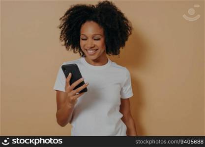 Happy African student smiling, using phone for positive news or chatting. Sand-colored wall background.