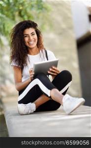 Happy African woman using digital tablet outdoors. Arab girl wearing sportswear and smiling in urban background
