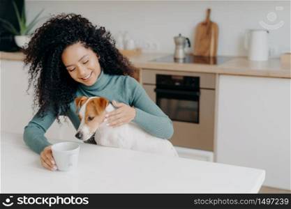 Happy Afro African woman with curly hairstyle treats dog in kitchen, pose at white table with mug of drink, enjoy domestic atmosphere, have breakfast together. People, animals, home concept.