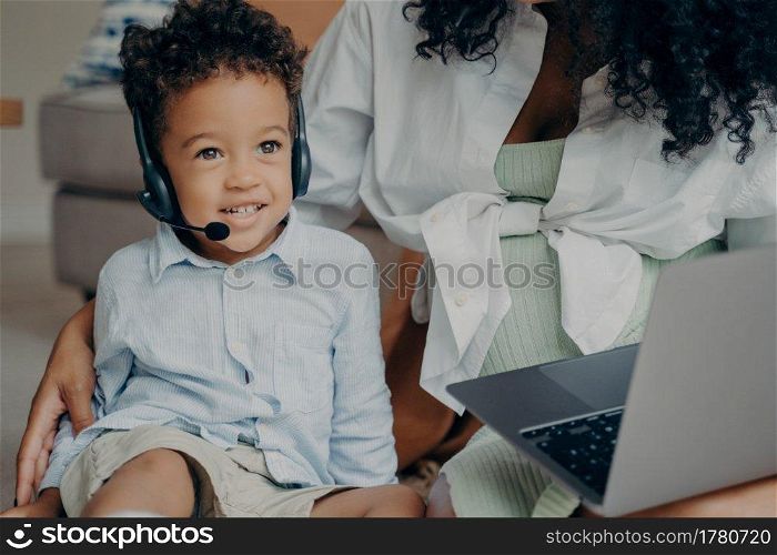 Happy afro american child with curly hair looking aside and smiling, wearing headset while having online meeting with dad, using laptop with his mother, boy talking with family through webcam. Happy afro american boy talking with family through webcam on laptop with mothers help