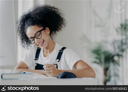 Happy Afro American female student writes down information, prepares for exams, sits in coworking space, has curly dark hair, wears optical glasses white t shirt and overalls, studies in spacious room