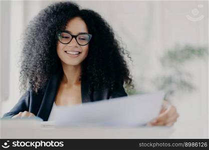 Happy Afro American woman with curly hairstyle, looks through documents, prepares financial report, wears transparent glasses and formal suit, poses in her cabinet, works on financial report