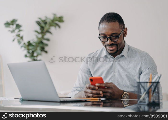 Happy afro businessman in glasses is texting on smartphone. Freelancer is working online on pc and having conversation with client at home office. Remote african american worker.. Remote african american worker in glasses is texting on smartphone at home office.