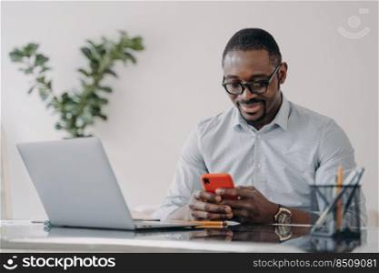 Happy afro businessman in glasses is texting on smartphone. Freelancer is working online on pc and having conversation with client at home office. Remote african american worker.. Remote african american worker in glasses is texting on smartphone at home office.
