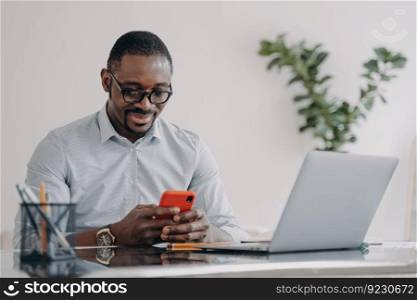 Happy afro businessman in glasses is texting on smartphone. Freelancer is working online on pc and having conversation with client at home office. Remote african american worker.. Remote african american worker in glasses is texting on smartphone at home office.