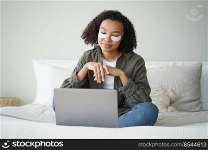 Happy afro girl applies eye patches and relaxing at home in weekend morning. Young hispanic woman having rest watching tv series. Skin moisturizing. Lovely curly girl is grooming herself in bedroom.. Happy afro girl applies eye patches and relaxing at home watching tv series. Weekend morning.