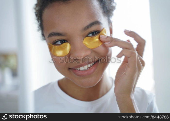 Happy afro girl is applying eye patches. Skin moisturizer with collagen serum. Beauty routine of lovely teenage african american girl. Mirror reflection of young spanish woman face. Morning skincare.. Happy afro teenage girl is applying eye patches. Skin moisturizer with collagen serum.