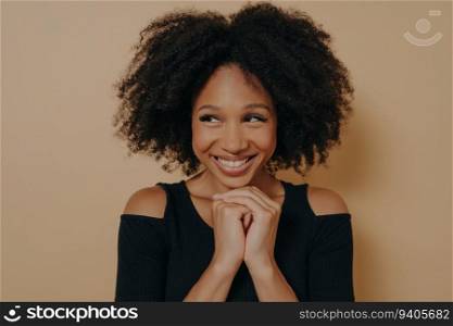 Happy Afro girl loves friends’ idea for next day, posing with hands under chin. Studio shot, dark beige wall background