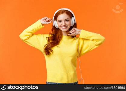 Happy and carefree excited redhead hipster girl like her new headphones, dancing with hands lifted up and smiling enjoy listening music over orange background, boost mood with favorite song.. Happy and carefree excited redhead hipster girl like her new headphones, dancing with hands lifted up and smiling enjoy listening music over orange background, boost mood with favorite song