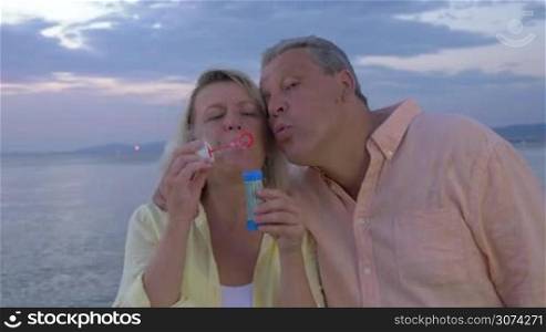 Happy and carefree senior man and woman blowing soap bubbles on the shore, sea and evening sky in background. Careless summer vacation