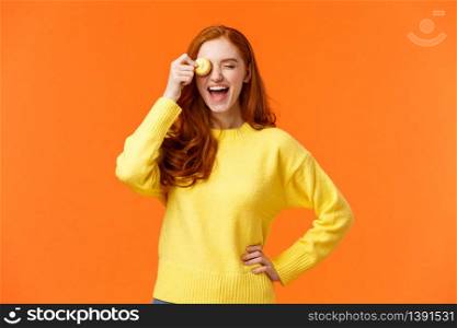 Happy and carefree smiling, laughing lovely redhead girl grinning, holding tasty macaron near eye, fooling around, having fun, eating delicious dessert at favorite cafe, orange background.. Happy and carefree smiling, laughing lovely redhead girl grinning, holding tasty macaron near eye, fooling around, having fun, eating delicious dessert at favorite cafe, orange background