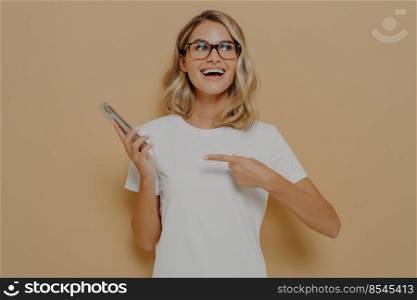 Happy and excited girl in spectacles pointing on mobile phone and looking aside with amazed face expression, reading unbelievable and good news in internet while posing in studio. Happy and excited girl in spectacles pointing on mobile phone and looking aside with amazed face expression