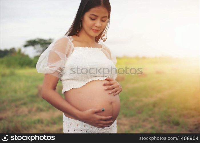 happy and proud pregnant Asian woman looking at her belly in a park at sunrise