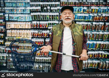 Happy angler holds net in fishing shop, hooks and baubles on background. Equipment and tools for fish catching and hunting, accessory choice on showcase in store, bait assortment. Happy angler holds net in fishing shop
