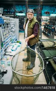 Happy angler holds net in fishing shop, hooks and baubles on background. Equipment and tools for fish catching and hunting, accessory choice on showcase in store, bait assortment. Happy angler holds net in fishing shop