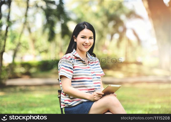Happy asia woman holding touch pad in hand / Beautiful young girl smiling looking tablet for entertainment and communication on park outdoors - woman student use technology education outside