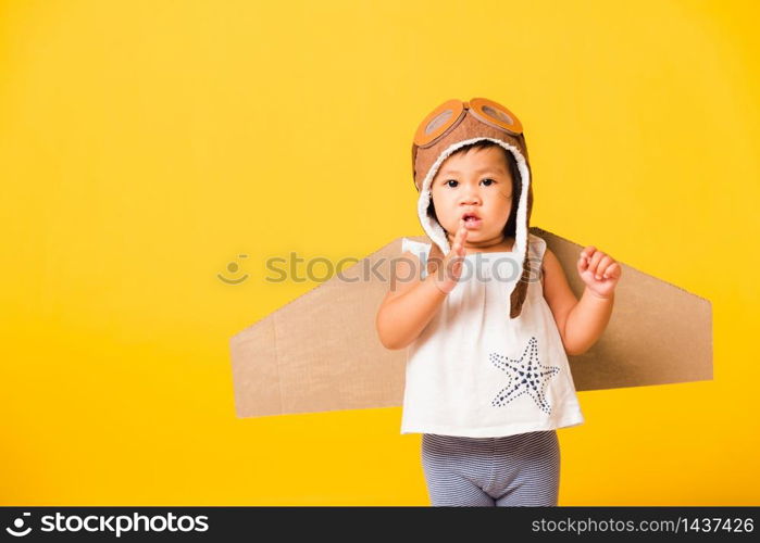 Happy Asian beautiful funny baby little girl smile wear pilot hat playing and goggles with toy cardboard airplane wings flying, studio shot isolated yellow background, Startup freedom concept