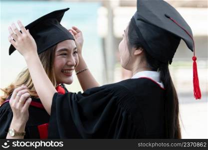 Happy Asian beautiful graduate student with University degree with black graduation cap with red tassels in dress is smiling while friend helping her get dress up. Concept of the graduation ceremony