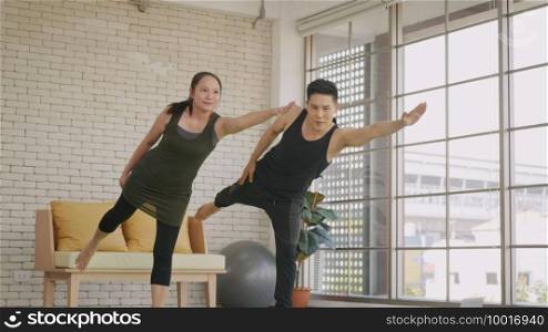 Happy Asian beautiful lifestyle family couple doing YOGA standing meditating on virabhadrasana 3 pose workout at home together on mat. Two people sport healthy concept