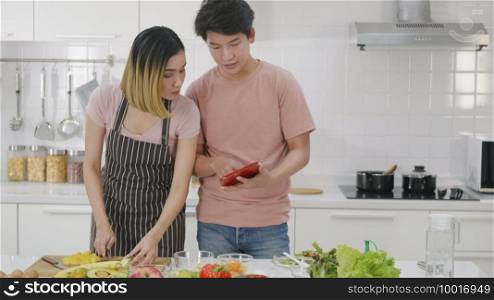 Happy Asian beautiful young family couple husband and wife cooking vegetable salad in kitchen together at home. The man and woman according online cooking class to recipe on laptop computer