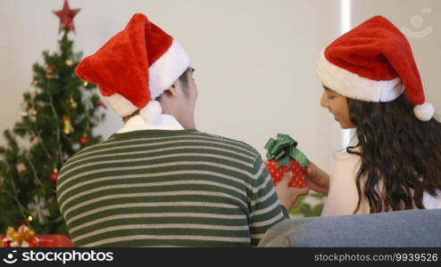 Happy Asian beautiful young family couple husband and wife sitting on sofa back view wearing a santa hat in front fireplace at home Christmas winter hugging giving gift box in living room,