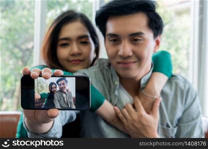 Happy Asian couple enjoy taking selfie photo with mobile phone in living room at home.