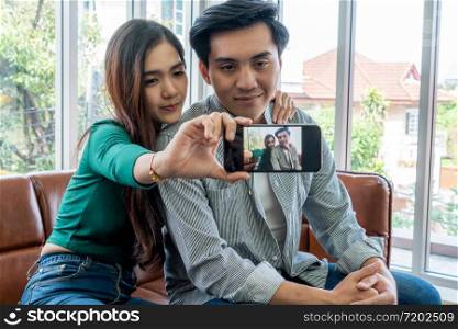 Happy Asian couple enjoy taking selfie photo with mobile phone in living room at home.