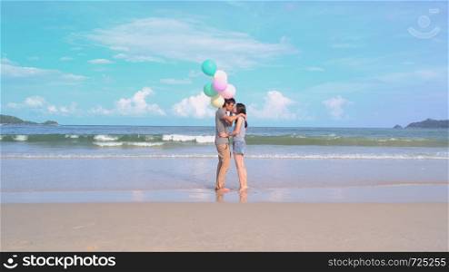 Happy Asian couple kissing, holding colorful balloons at the beach during travel trip on holidays vacation outdoors at ocean or nature sea at noon, Phuket, Thailand
