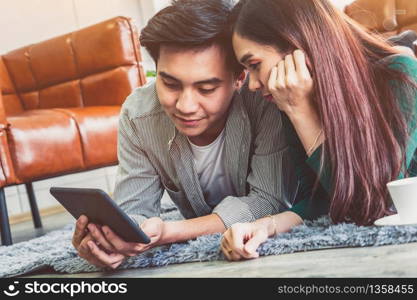 Happy Asian couple use tablet while lying down on carpet at living room floor. Love and relationship concept.