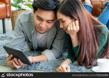Happy Asian couple use tablet while lying down on carpet at living room floor. Love and relationship concept.