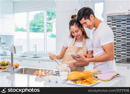 Happy Asian couples cooking and baking cake together in kitchen room. Man and woman looking to tablet follow recipe step at home. Love and happiness concept. Sweet honeymoon and Valentine day theme