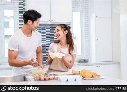 Happy Asian couples cooking and baking cake together in kitchen room. Man and woman looking to tablet follow recipe step at home. Love and happiness concept. Sweet honeymoon and Valentine day theme