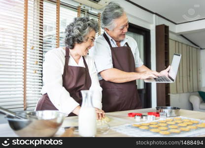 Happy Asian Elderly couple in brown aprons using laptop computer for learning online cookies class while preparing dough together. Grandfather and Grandmother having fun in a kitchen.